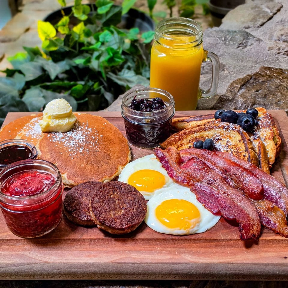 Breakfast board with pancakes, French toast, eggs, bacon, sausage, and orange juice.